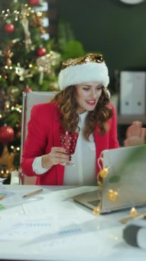 Noel zamanı. Gülümseyen şık, küçük işletme sahibi, Noel Baba şapkalı, kırmızı ceketli, dizüstü bilgisayarı ve şarap kadehiyle modern yeşil ofiste Noel ağacı olan bir kadın..