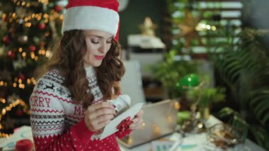 Noel zamanı. Noel Baba şapkası ve kırmızı Noel süveteri içinde gülümseyen şık bir iş kadını. Zarfla ve parayla modern yeşil ofiste Noel ağacı ile..