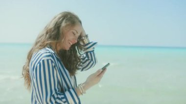 Deniz kıyısında akıllı telefonuyla gülümseyen şık bir kadın..