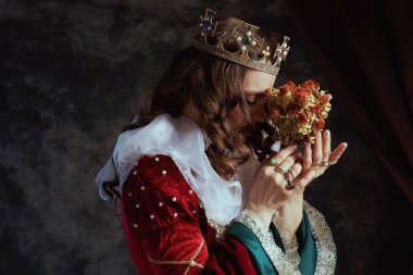 Kurumuş çiçekli kırmızı elbiseli ortaçağ kraliçesi, koyu gri arka planda beyaz yaka ve taç..