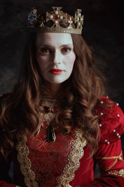 Kırmızı elbiseli, beyaz makyajlı ve tacı koyu gri arka planda olan ortaçağ kraliçesi..