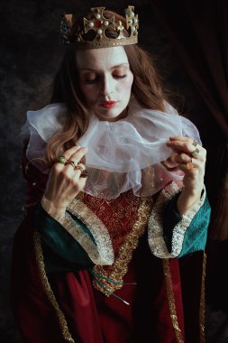 Kırmızı elbiseli, beyaz yakalı ve tacı koyu gri arka planlı ortaçağ kraliçesi..