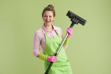 Bahar temizliği. Yeşil önlüklü mutlu orta yaşlı kadın ve yeşil elektrikli süpürgeye izole edilmiş lastik eldivenler..