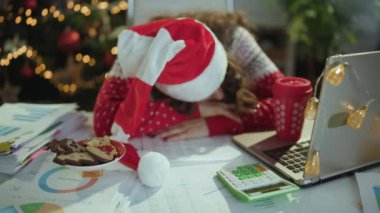 Noel zamanı. Noel Baba şapkalı, kırmızı Noel süveterli, belgeli, laptoplu, modern yeşil ofiste Noel ağacıyla uyuyan orta yaşlı, şık bir iş kadını..