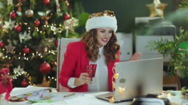 Noel zamanı. 40 yaşında, Noel Baba şapkalı, kırmızı ceketli, dizüstü bilgisayarı ve şarap kadehiyle modern yeşil ofisinde Noel ağacı olan bir iş kadını portresi..