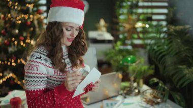 Noel zamanı. Düşünceli modern 40 yaşında Noel Baba şapkalı küçük işletme sahibi kadın ve kırmızı Noel süveteri içinde zarf ve parayla modern yeşil ofis ve Noel ağacı..