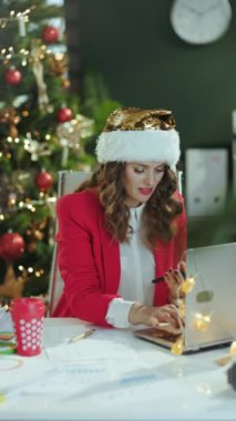 Noel zamanı. Kırmızı ceketli, belgeli, dizüstü bilgisayarlı, akıllı telefon uygulamalı, modern yeşil ofisteki Noel Baba şapkalı, Noel ağacıyla çalışan bir gülümseme..