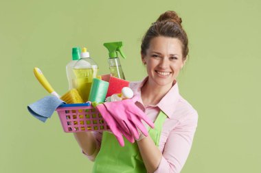 Bahar temizliği. Yeşil önlüklü mutlu orta yaşlı ev kadını ve yeşil üzerine izole edilmiş lastik eldiven ve temizlik ürünleri sepeti..