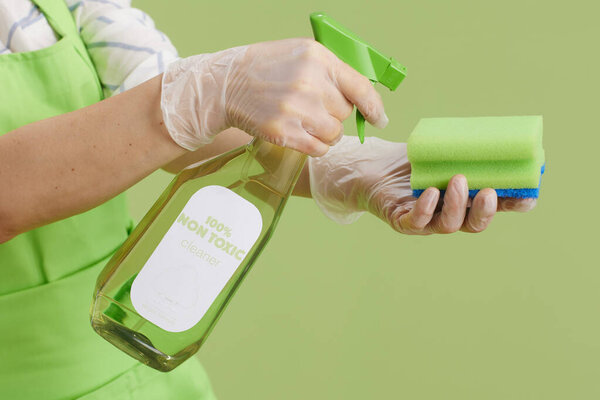 Весенняя уборка. Крупный план современной женщины в зеленом фартуке и резиновых перчатках, изолированных на зеленом с бутылкой органического чистящего средства и чистящей губки.