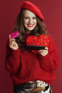 Happy Valentine. smiling elegant woman in red sweater and beret with red heart, smartphone and credit card making online shopping on e-commerce site.