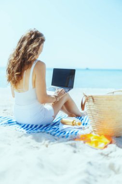 Saman çantası, dizüstü bilgisayarı boş ekranı ve çizgili havlusuyla deniz kıyısındaki modern kadının arkasından görüldü..