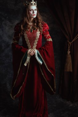 Kırmızı elbiseli, beyaz makyajlı ve tacı koyu gri arka planda olan ortaçağ kraliçesi..