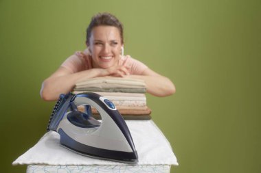 Gülümseyen bir kadın, önünde ütüyle katlanmış bir elbisenin üzerine uzanır ve soluk zeytin yeşili bir arka plana yaslanır. Rahatlamış ve mutlu görünüyor..