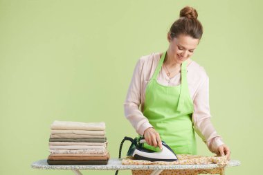 Saçları topuz şeklinde sırıtan bir kadın desenli kumaş ütülüyor. Soluk yeşil arka planda pembe bir gömlek ve yeşil bir önlük giyiyor. Düzgünce katlanmış bir kıyafet yığını yakınlarda duruyor..