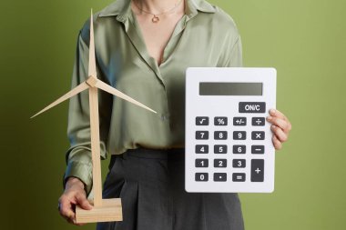 Açık yeşil zemine karşı ahşap bir rüzgar türbini modeli ve hesap makinesi tutan kadın çevresel farkındalığı ve finansal sürdürülebilirliği sembolize ediyor..
