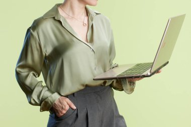 Açık yeşil bluz ve gri pantolon giyen kendine güvenen bir kadın, elinde parlak yeşil bir arkaplana karşı dizüstü bilgisayar tutuyor. Bu da modern ve verimli bir çalışma ortamı olduğunu gösteriyor..