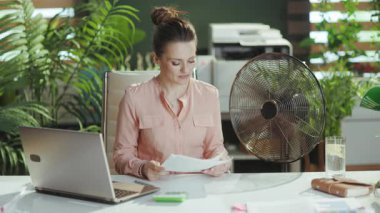 Sürdürülebilir iş yeri. Modern yeşil ofiste elektrikli vantilatör ve dizüstü bilgisayarıyla gülümseyen orta yaşlı bir muhasebeci kadın..