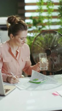 Sürdürülebilir iş yeri. Gülümseyen 40 yıllık modern kadın işçi modern yeşil ofiste elektrikli yelpaze, su ve limon ile.