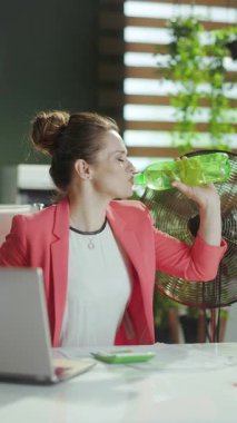 Sürdürülebilir iş yeri. Modern yeşil ofisteki 40 yaşındaki mutlu iş kadını kırmızı ceketli, su ve elektrikli vantilatörlü..