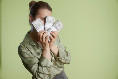 40 'lı yaşlarda bir kadın 100 dolarlık banknotlar tutuyor, yüzünü kısmen gizliyor, açık yeşil bir fon karşısında servet ve maddi kazanç öneriyor..