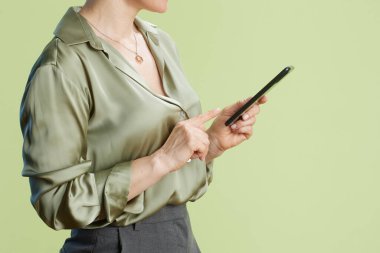 40 'lı yaşlarda bir kadın açık yeşil saten bluz ve gri etek giyen akıllı telefon ekranına dokunuyor. Basit bir yeşil zemin onun profesyonel davranışlarını ve modern teknoloji kullanımını vurguluyor..