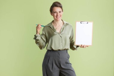 30 'lu yaşlarda gülümseyen bir kadın açık yeşil arka planda elinde bir pano ve kalem tutuyor. İpeksi yeşil bir bluz ve gri bir pantolon giyiyor..