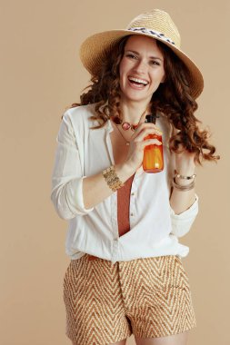 Plaj tatili Beyaz bluz ve şortla gülümseyen stil sahibi kadın saç bakım ürünleri ve yaz şapkası ile bej arka planda izole edilmiş..