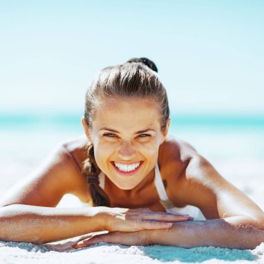 Saçları topuz şeklinde genç bir kadın kumlu bir plajda uzanmış kameraya gülümsüyor. Parlak mavi gökyüzü ve açık mavi okyanus neşe dolu, tasasız havayı artırır..