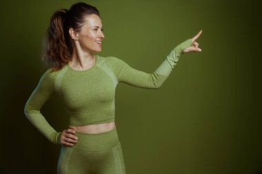 30 'lu yaşlarda canlı bir kadın, yeşil atletik kıyafetler giymiş, gülümseyen ve puanlı, enerjik, zinde ve aktif bir yaşam tarzını yeşil bir zemine karşı teşvik eden.