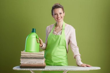 Gülümseyen bir kadın ütü masasında katlanmış giysilerin yanında deterjan şişesi tutuyor. Pembe gömlek, yeşil önlük, zeytin yeşili arka plan. Çamaşır, temizlik..