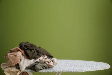 Toprak tonlu bir yığın, zeytin yeşili bir arka planda ütü masasında gelişigüzel duruyor. Yumuşak ışık, sakin ve evcil bir atmosfer olduğunu gösteriyor..