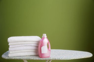 Beyaz havlular ve pembe bir deterjan şişesi bir ütü masasında duruyor. Hepsi de zeytin yeşili bir zemine karşı. Temiz ve düzenli estetik..