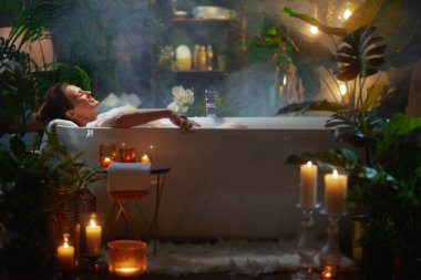 Bir kadın yeşil bitkiler ve mum ışığı ile çevrili, gözleri kapalı köpük banyosunda dinlenir. Buhar yükselir, huzurlu, spa benzeri bir atmosfer yaratarak nihai rahatlama için.