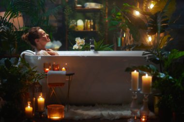 Bir kadın, yemyeşil ve yumuşak mum ışığıyla çevrili beyaz bir küvette rahatlar. Huzuru ve kendine bakımı teşvik eden sıcak ve rahatlatıcı bir atmosfer yaratır..