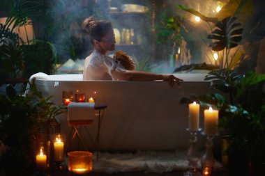 Köpük banyosundaki bir kadın omzunu süngerle ovalıyor. Etrafı mum ve bitkilerle çevrili. Rahatlatıcı ve lüks bir spa deneyimi..