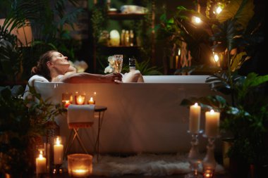 Bir kadın beyaz bir küvette şampanyayla, bitkilerle ve mum ışığıyla rahatlıyor. Sahne huzurlu bir spa deneyimi yaratıyor..