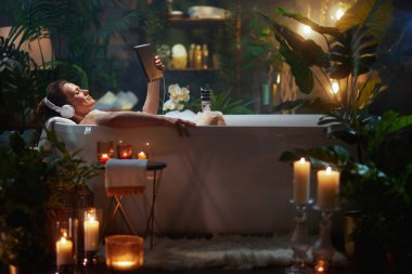 Orta yaşlı bir kadın elinde tablet ve kulaklıkla köpük banyosunda dinleniyor. Etrafı bitkiler ve mum ışığıyla çevrili. Lüks bir kişisel bakım anı..