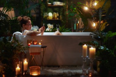 Beyaz bir küvette dinlenen bir kadın, bitkiler ve mumlarla dolu bir banyoda cep telefonuna bakıyor. Olay yeri kendine özen ve huzur dolu bir rahatlığı çağrıştırıyor..