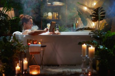 Yeşil bitkiler ve mum ışığıyla çevrili köpük banyosunda gülümseyen bir kadın. Bir rahatlama, kişisel bakım ve dijital rahatlama anı..