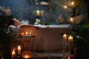 Bir kadın yemyeşil ve yumuşak mum ışığıyla çevrili kabarcıklarla dolu bir küvette rahatlar. Spa benzeri bir ortam rahatlamayı, kendine bakmayı ve huzurlu bir lüksü teşvik eder..