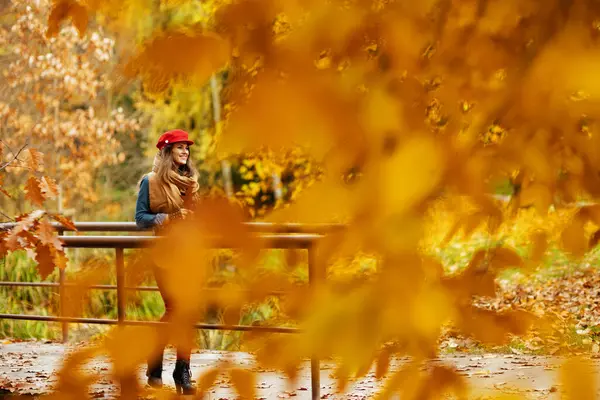 Merhaba sonbahar. Kot gömlekli, kırmızı şapkalı, güz yaprağı ve eşarplı mutlu şık kadın şehir parkında yürüyor..