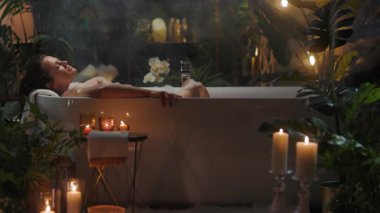 Bir kadın, bitkiler ve mumlarla çevrili bir köpük banyosunda dinleniyor, loş bir banyoda buhar yükseliyor. Rahatlama ve kendine bakımı teşvik eden sakin bir spa sahnesi..