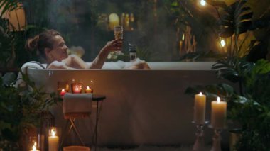 Bir kadın, şampanyası ve yemyeşil ışıklarla çevrili rahatlatıcı köpük banyosundan zevk alır. Huzurlu, spa benzeri bir atmosfer şımartmayı ve kendini korumayı teşvik eder..
