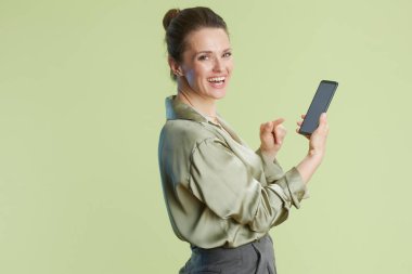 Gülümseyen bir kadın akıllı telefonu işaret ederek açık yeşil bir arka plana karşı heyecan ve güveni ifade eder. Kıyafeti profesyonel bir ortam olduğunu gösteriyor..