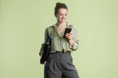 Kadın elinde evrak çantası tutarken akıllı telefonuna bakıyor. Açık yeşil düğmeli bluz ve gri pantolon yumuşak yeşil arka plana karşı profesyonel bir ayar önerir.