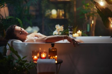 Orta yaşlı bir kadın lüks bir köpük banyosunda dinleniyor, gözleri huzur içinde dinleniyor. Mumlar, dekoratif bir şişe, ve yumuşak ipli yapraklar sakin, spa benzeri bir dinlenme yeri oluşturur..