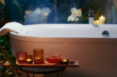 Beyaz bir küvet dinlendirici bir sağlık inzivası için hazırlandı. Buhar yükseliyor ve ahşap tepside kehribar yanığı mumlar ve banyo malzemeleri bulunuyor. Bulanık yeşil bitkiler ve orkideler mükemmel bir akşam için rahat ve rahatlatıcı atmosferi tamamlıyorlar..