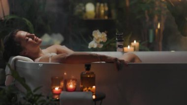 30 'lu yaşlarda bir kadın lüks bir köpük banyosunda huzur ve şımarıklık bulur. Etrafı yemyeşil bitkiler, mumlar ve güzellik ürünleriyle çevrilidir. Huzurlu ve huzurlu bir kaplıca kaçışı yaratır..