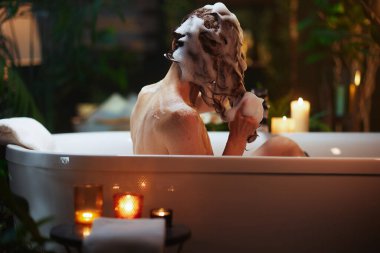 Arkasından görülen dingin bir kadın köpükle kaplı saçlarını titreyen mumlar ve yemyeşil bitkilerle çevrili beyaz bir küvette yıkıyor. Bu samimi sahne, evdeki sükuneti ve lüks bir kaplıca deneyimini anımsatıyor..