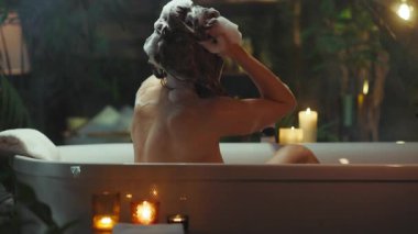 Arkadan görülen bir kadın, omuz hizasında kahverengi saçlarını beyaz bir küvette şampuanla yıkıyor. Yanıp sönen mumlar, sicim ışıkları ve ev bitkileriyle çevrili sahne huzur ve özgüven yayıyor..